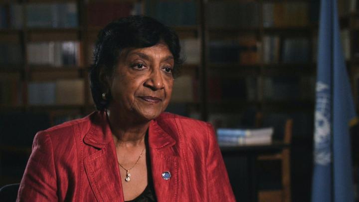 Judge Pillay  - a woman with brown skin and short dark hair sits in front of a bookshelf, wearing a red jacket over a black top, pearl earrings and a gold necklace. A blue United Nations flag is visible in the background on the right. She is looking slightly to her left with a composed, thoughtful expression. The lighting focuses on her face, while the background remains softly blurred.