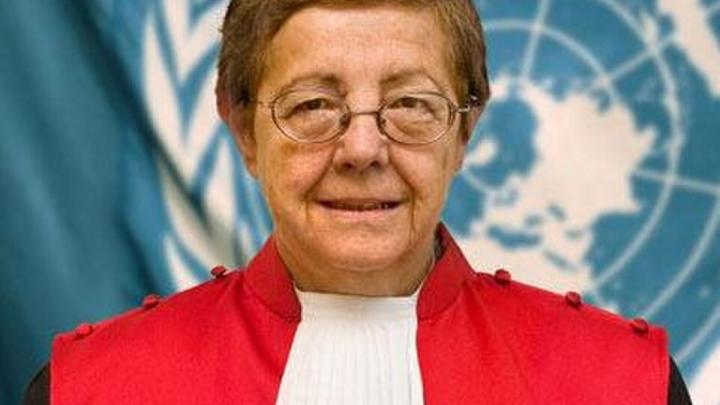 The photo shows a woman with short, light brown hair and oval wire-rimmed glasses, smiling at the camera. She's wearing formal judicial robes: a red robe with a stiff white jabot (the pleated white collar piece worn by judges) at the front, and black sleeves visible on the shoulders. The robe has decorative buttons along the shoulder seams.  Behind her is a large United Nations flag — pale blue with the white UN emblem (olive branches surrounding a world map) visible over her right shoulder. The setting and