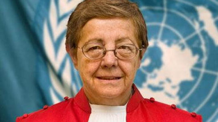 A formal portrait of an older woman facing the camera and looking directly toward the viewer. She has short, neatly combed brown hair, wears thin oval eyeglasses, and has a calm, slight smile. She is dressed in a bright red formal jacket with decorative shoulder details, over a white high-neck blouse.  Behind her is a large blue-and-white United Nations emblem and flag imagery, suggesting an official UN setting. The photograph is closely framed from about the chest upward, with the woman cent