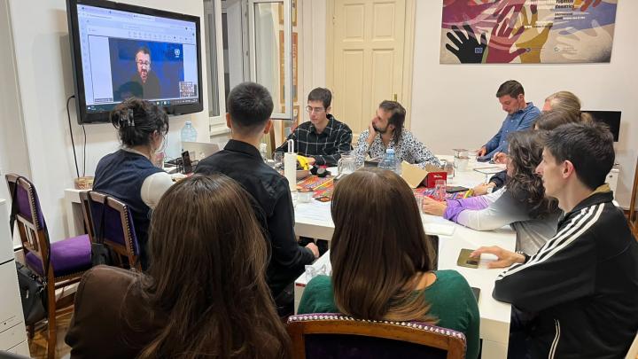 Ten people sit around an oval table, facing a wall-mounted TV displaying a blue presentation slide. The group appears engaged in a meeting or class within a neutrally decorated room with framed artwork on the walls.